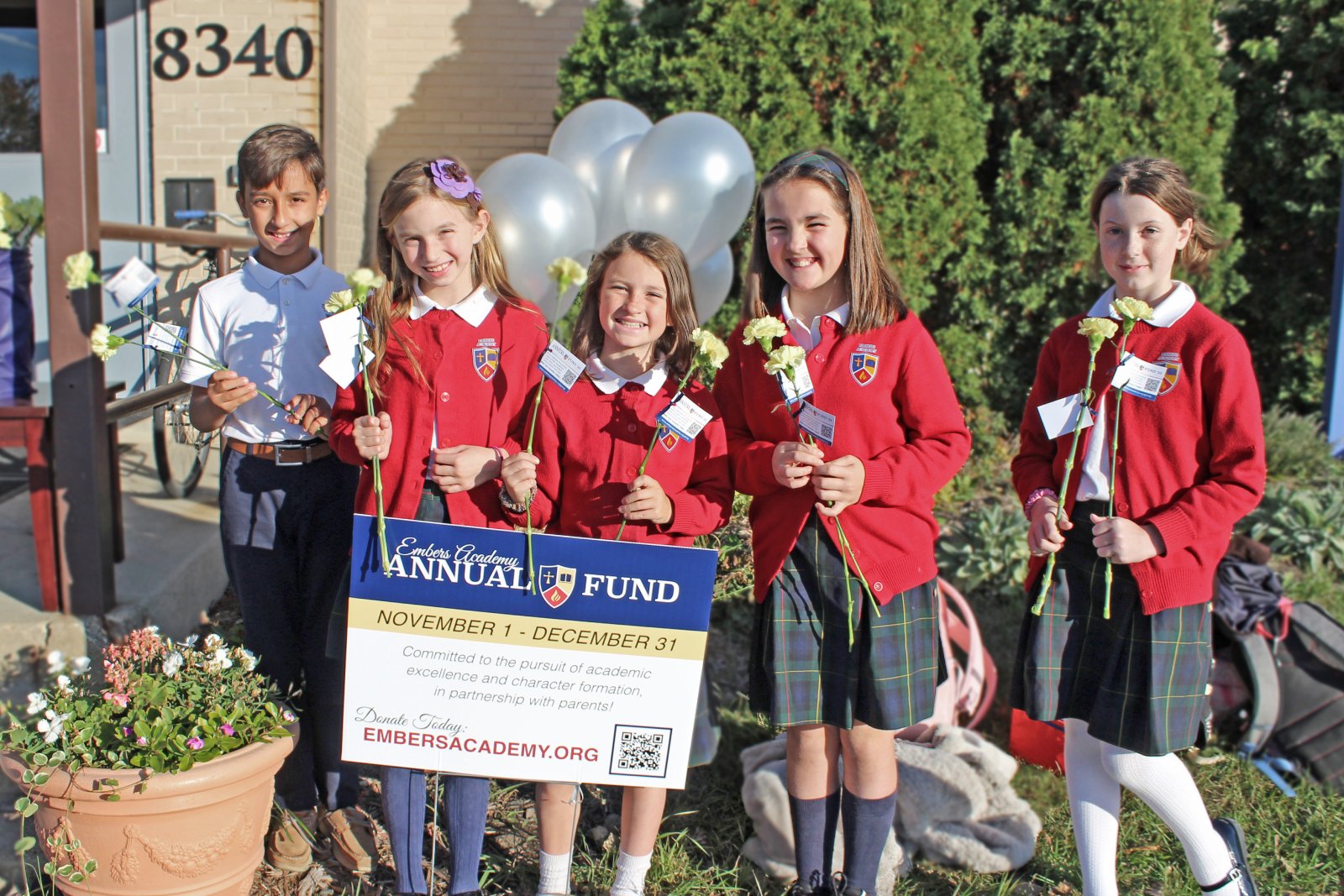 Five smiling students in red cardigans and plaid skirts pose outdoors, holding carnations and a fundraising sign for Embers Academy Annual Fund.